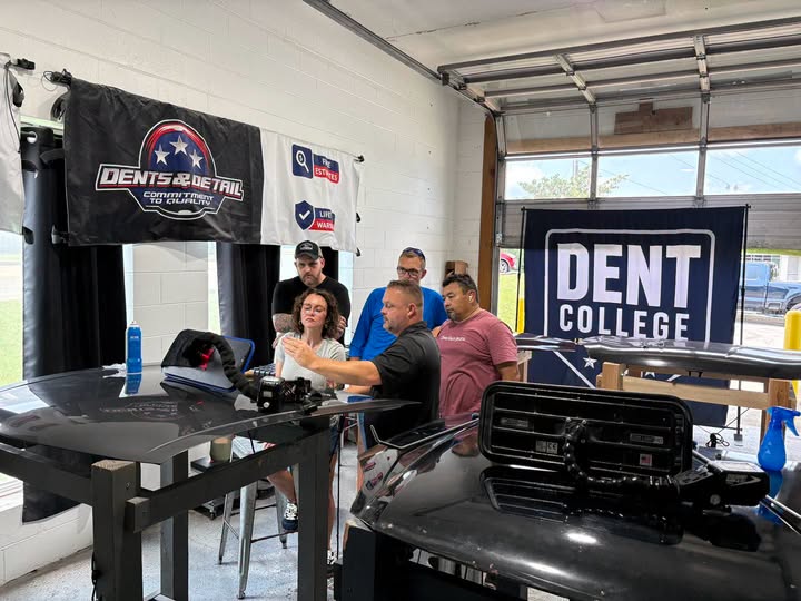 A Dent College instructor leading a small hands-on paintless dent repair class, demonstrating dent removal on a vehicle panel while students watch closely